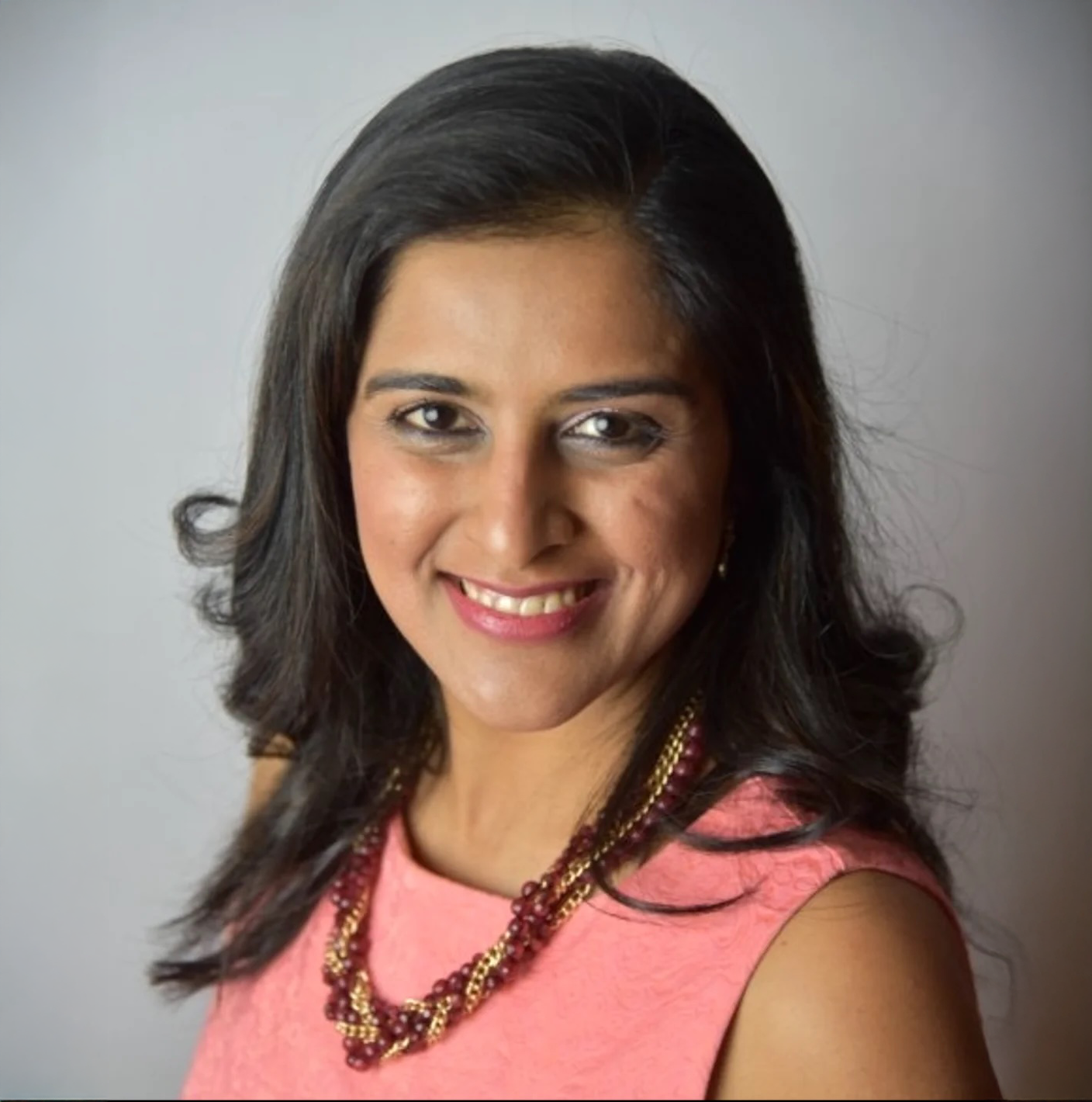 A headshot of Sadaf Sajwani smiling. She has dark hair and a coral-colored top, wearing a beaded necklace.