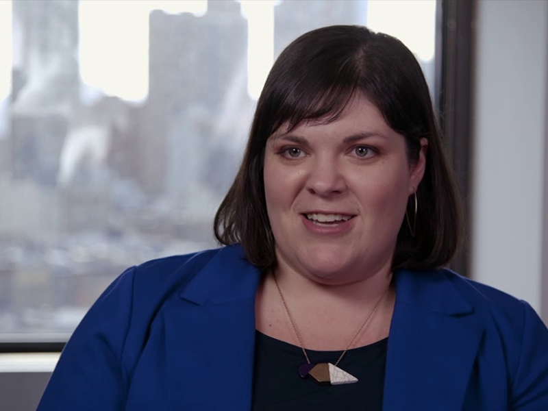 A woman in a blue suit gives an interview while sitting in front of a window that looks out onto a city.